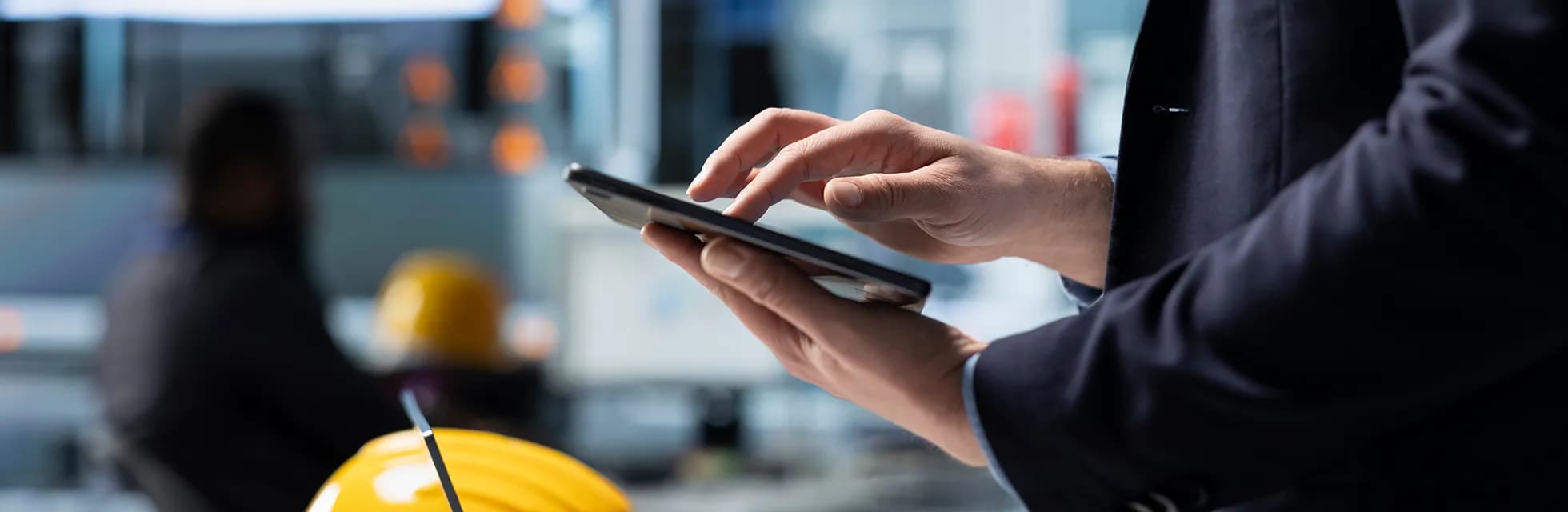 Detailný záber na muža v obleku používajúceho tablet s rozostreným pozadím priemyselného pracoviska a žltou ochrannou prilbou v popredí.