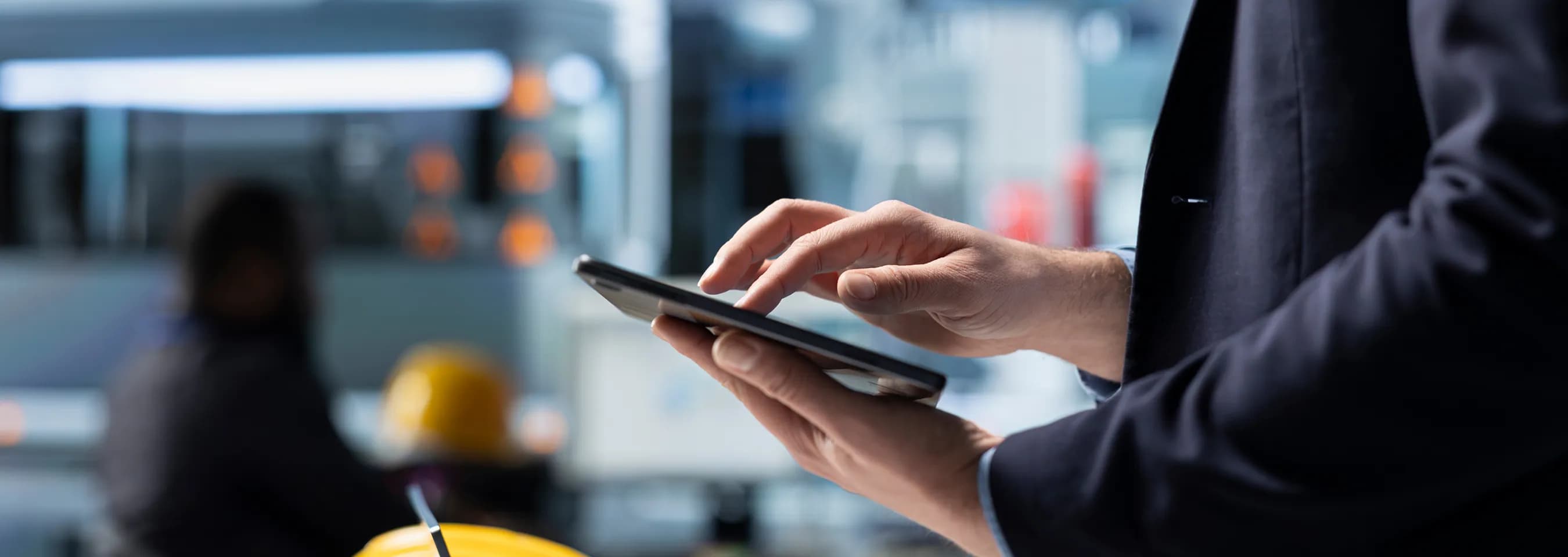 Detailný záber na muža v obleku používajúceho tablet s rozostreným pozadím priemyselného pracoviska a žltou ochrannou prilbou v popredí.
